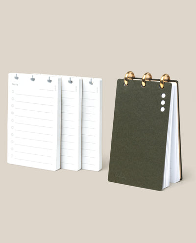 The Discbound Analog + 3 Refills, featuring a compact dark green ring-bound notepad with brass discs, stands upright beside three stacks of daily planner refill pages on a beige background.