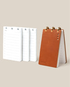 The Leather Discbound Analog + 3 Refills (Tan) features a tan full-grain leather notepad with brass rings and three stacks of white daily planner refills, all displayed on a beige background.