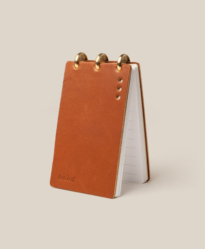 The Leather Discbound Analog (Tan) is a small notepad with three brass rings, a full-grain leather cover in brown featuring four metal studs and "Analog" embossed on the corner. Lined pages provide ideal organization.
