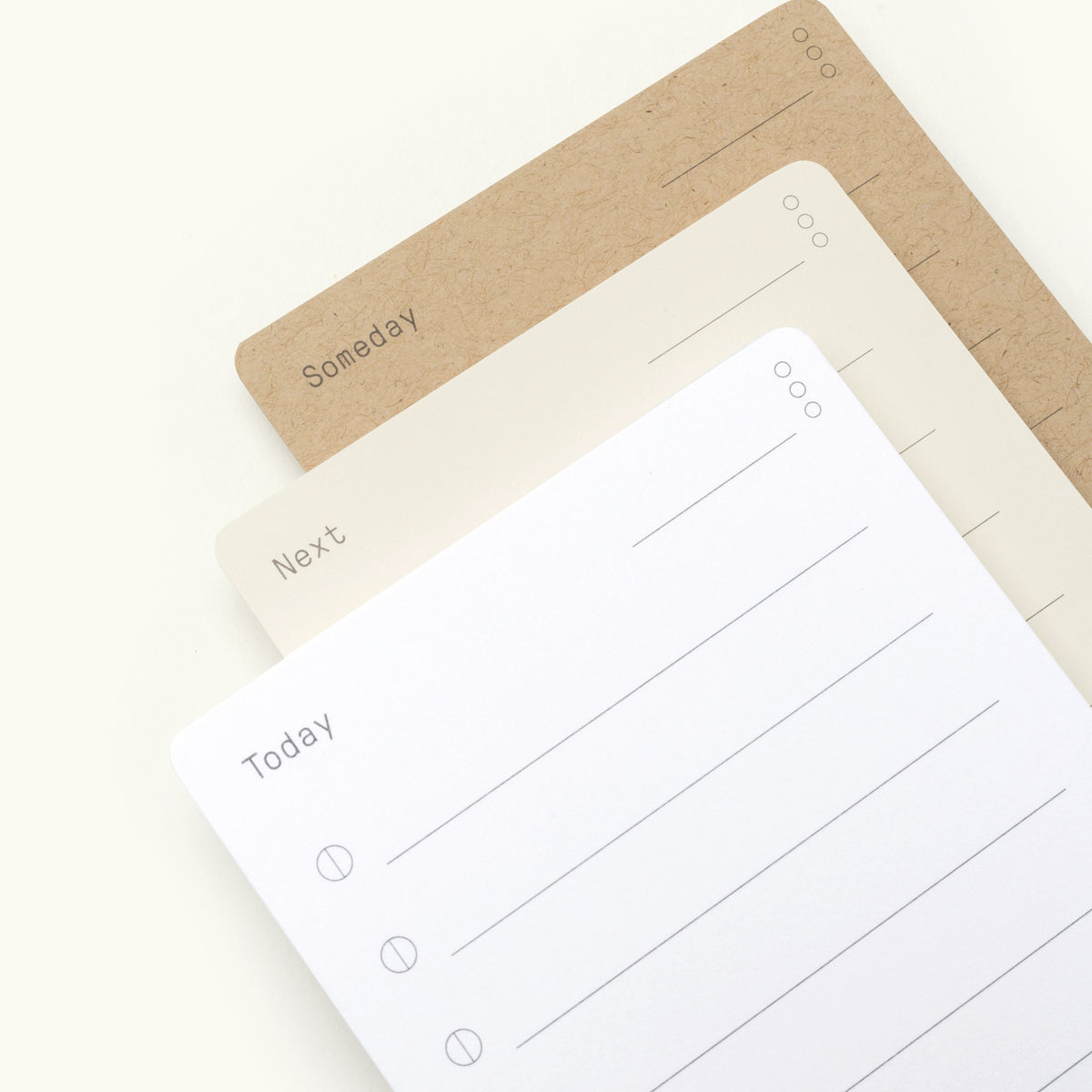 Three notepads are stacked, labeled Today (white), Next (off-white), and Someday (brown), each with lines and circles for making lists. The background is a light, neutral color.
