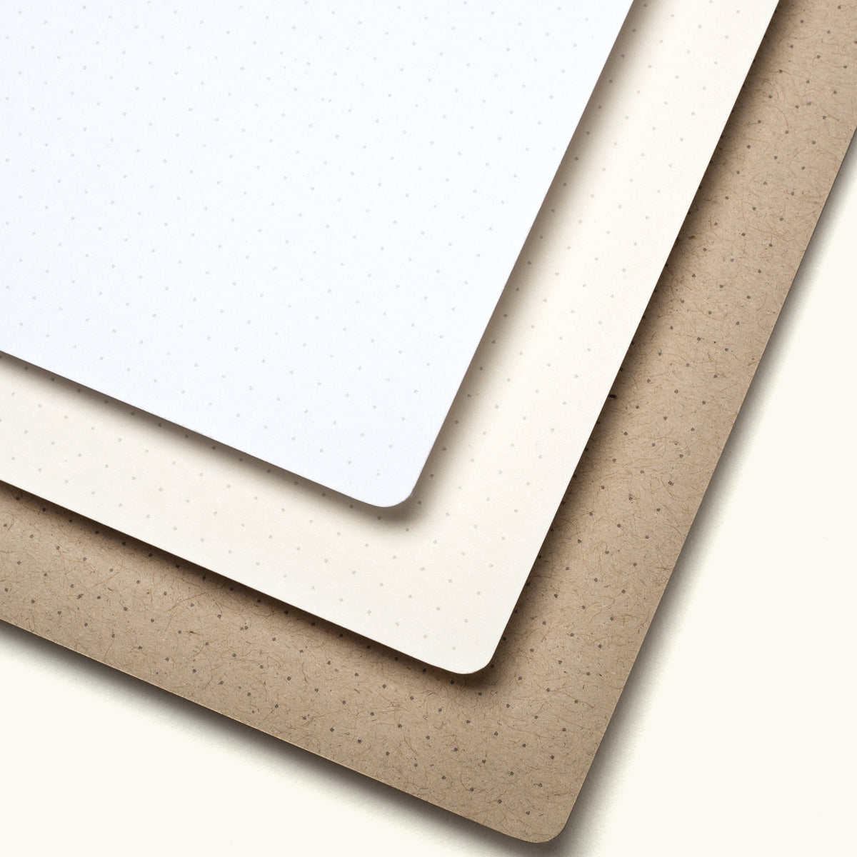 Three sheets of paper stacked diagonally: a white dotted sheet on top, a cream-colored sheet in the middle, and a brown kraft paper sheet on the bottom, all with smooth, rounded corners.