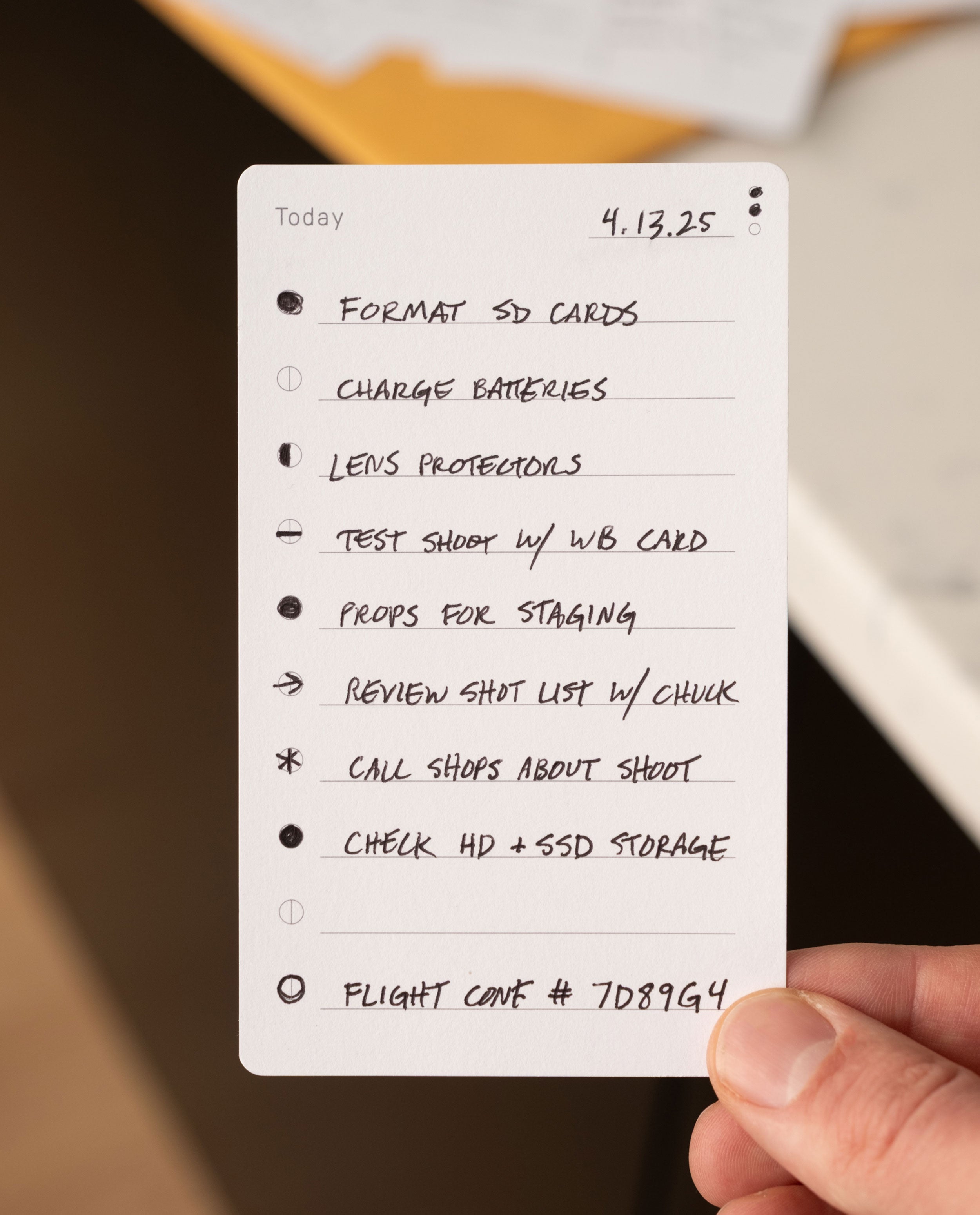 A hand holds a handwritten checklist titled Today (4.13.25) on Analog Trial Pack card stock, listing photography prep steps like formatting SD cards, charging batteries, reviewing a shot list, and noting a flight case number at the bottom.