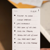 A hand holds a checklist dated 4.13.25 with tasks including formatting SD cards, charging batteries, lens protection, test shoot, staging props, reviewing a shot list, calling shops, checking storage, and a flight code.