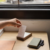 A hand pulls a white Analog - Today Card (3-Pack) from a wooden box on a desk. Nearby are a closed notebook and green power bank on dark fabric. The 3x5 card has text, with a window visible in the background.