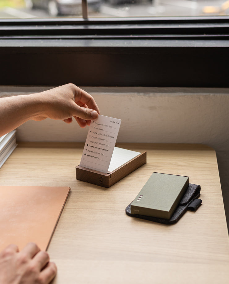 A hand pulls a white Analog - Today Card (3-Pack) from a wooden box on a desk. Nearby are a closed notebook and green power bank on dark fabric. The 3x5 card has text, with a window visible in the background.