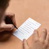 Using a black pen, someone writes tasks like “Follow up with Jim” and “Schedule meeting” on a small, white 3x5 card from the Analog Trial Pack, placing the premium card stock on a light brown surface.