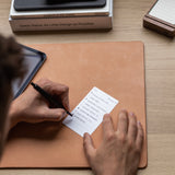 Someone writes a to-do list on an Analog Annual Refill card while sitting at a desk with a leather mat, tablet, and stacked books, including Dieter Rams: As Little Design as Possible.