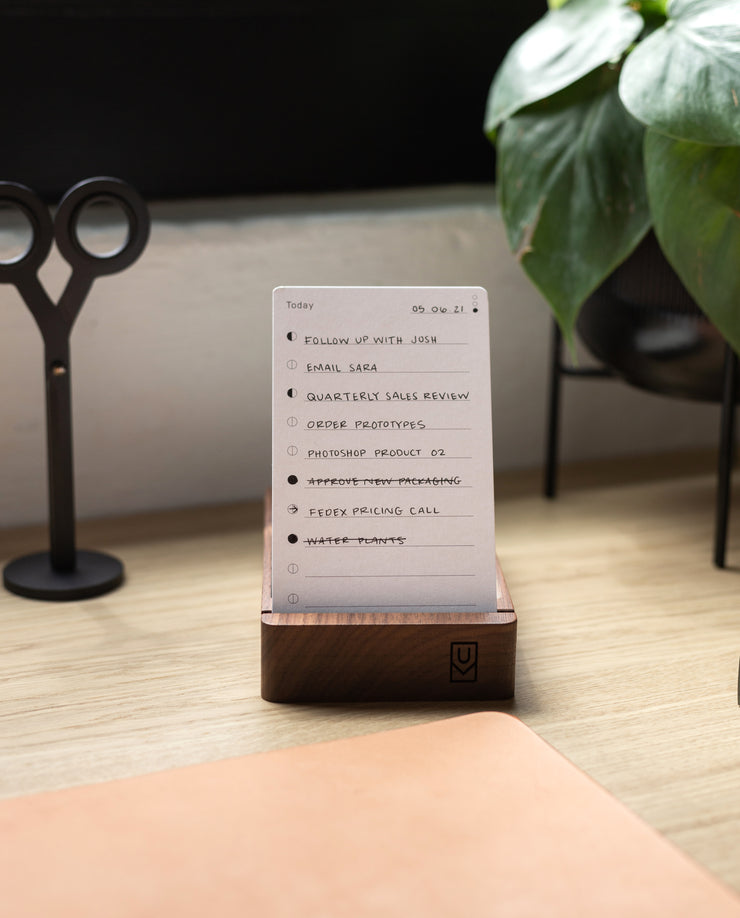 Analog - Today Cards (3-Pack), featuring a dot grid and to-do list tasks like "Email Sara" and "Order prototypes," rest on a wooden stand atop a desk with scissors, a plant, and part of a tan desk pad also in view.