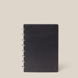 The Discbound Heirloom Journal (Black Leather - Standard 6" x 8.5") features a refillable design with a full-grain black leather cover and metal rings on the left, shown standing upright against a light beige background.