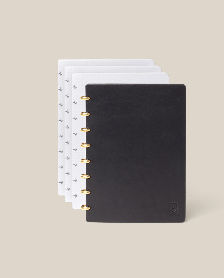 The Discbound Heirloom Journal Bundle (Black Leather - Standard 6" x 8.5" + 3 Refills) is open with gold rings, showing dotted and patterned pages fanned out behind the cover on a light beige background.
