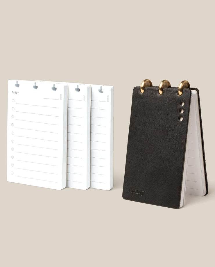 The Leather Discbound Analog + 3 Refills (Black), a compact black full-grain leather notepad with gold rings, stands upright beside three stacks of loose lined refill cards on a light beige background.