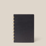 The Discbound Heirloom Journal (Black Leather - Standard 6" x 8.5") features a smooth black cover and gold disc binding, elegantly displayed upright against a beige background—a stylish refillable notebook for daily notes.