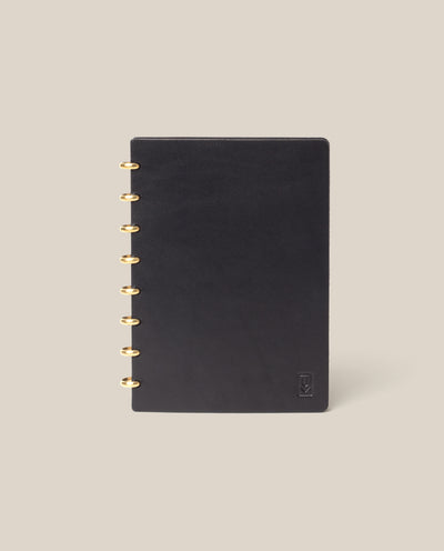 The Discbound Heirloom Journal (Black Leather - Standard 6" x 8.5") features a smooth black cover and gold disc binding, elegantly displayed upright against a beige background—a stylish refillable notebook for daily notes.