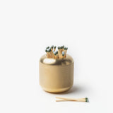 The Craighill Match Striker (Brass) holds strike-anywhere matches upright in its modern, minimalist solid brass container with a textured surface. Two matches rest beside it on a white background.