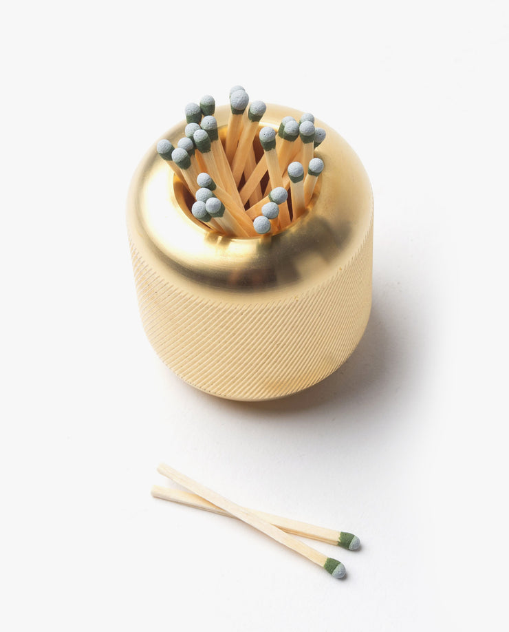 The Craighill Match Striker (Brass) is a gold, round solid brass holder filled with wooden matches, with three strike anywhere matches placed beside it on a white surface.