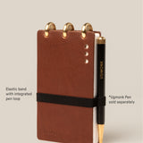 A small brown Leather Discbound Analog notebook with full-grain leather covers, gold rings, and a black elastic band with pen loop. A black and gold Ugmonk pen is beside it; the pen is sold separately.
