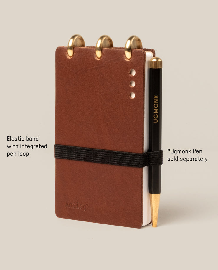 A small brown Leather Discbound Analog notebook with full-grain leather covers, gold rings, and a black elastic band with pen loop. A black and gold Ugmonk pen is beside it; the pen is sold separately.