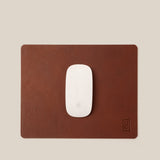 A white Apple Magic Mouse rests on a rectangular Ugmonk Leather Mousepad (Brown) featuring a small embossed logo in the bottom right corner, set against a beige background.