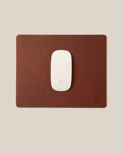 A white Apple Magic Mouse rests on a rectangular Ugmonk Leather Mousepad (Brown) featuring a small embossed logo in the bottom right corner, set against a beige background.