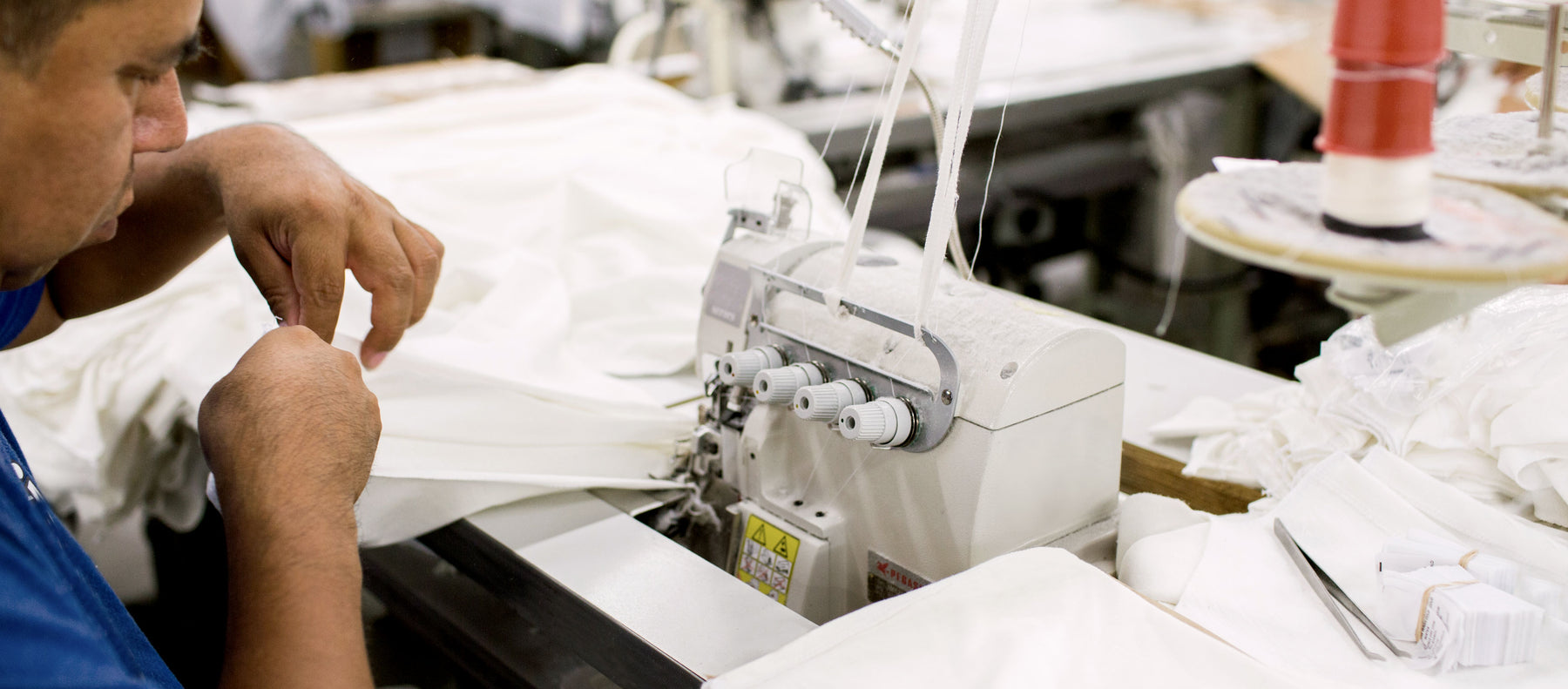 A person uses a sewing machine to stitch white fabric in a textile factory, with various threads and materials visible on the worktable.