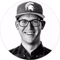 A smiling person wearing glasses, a button-up shirt, and a cap with a Spartan helmet logo, seen in a black-and-white, circular portrait.