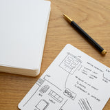 A black pen rests on a wooden desk beside an open notebook and a sheet of Analog Dot Grid Cards - Large (1-Pack) covered with sketches, layout diagrams, and handwritten notes.