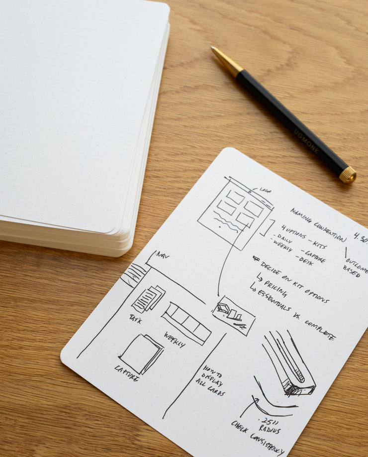 A black pen rests on a wooden desk beside an open notebook and a sheet of Analog Dot Grid Cards - Large (1-Pack) covered with sketches, layout diagrams, and handwritten notes.