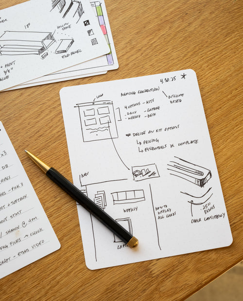 A black pen rests on a notepad with hand-drawn project diagrams, alongside Analog Dot Grid Cards - Large (12-Pack) and 100lb card stock, all arranged on a wooden desk scattered with notes and papers.
