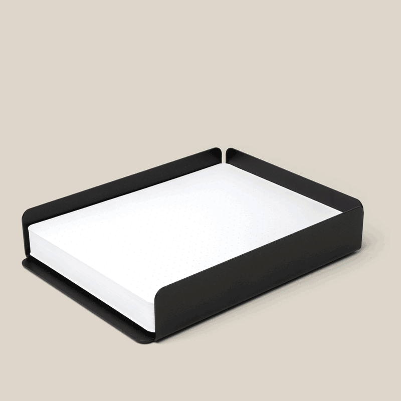 A black metal paper tray holds a stack of white paper on a beige surface. The tray has minimalist, rounded edges and an open front for easy access to the paper.