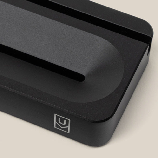 A close-up of a matte black, rectangular metal object with a shallow groove and rounded edge, featuring a white geometric logo on the corner, set against a light beige background.