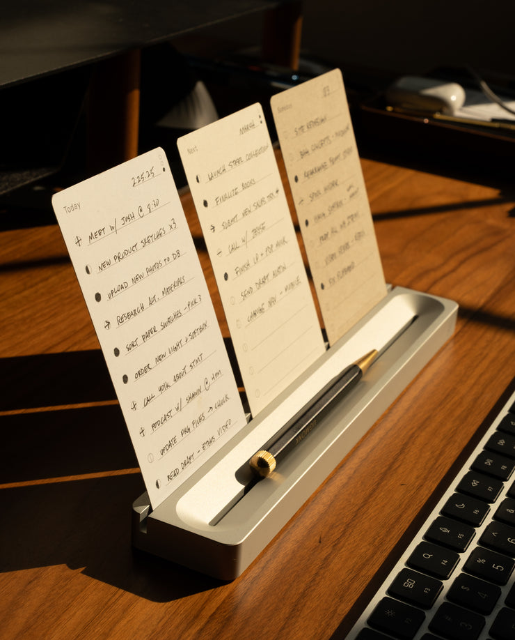 A desk organizer holding three handwritten to-do lists and a pen sits on a wooden desk beside a computer keyboard, bathed in warm sunlight.