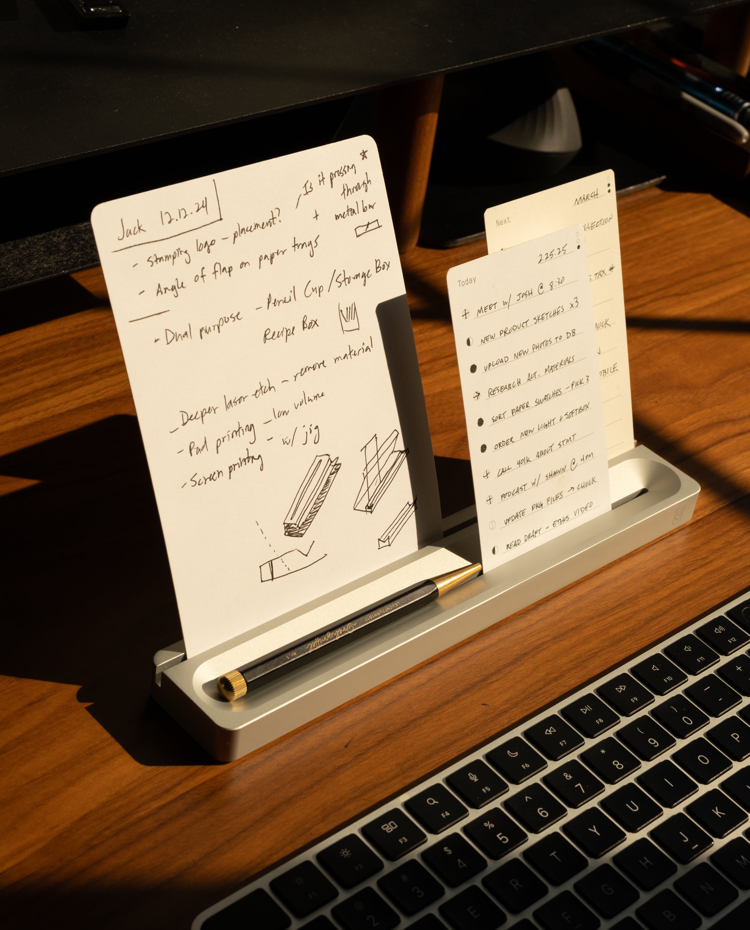 Two handwritten note cards from the Analog Card Bar - Essentials Kit (Silver Aluminum) sit in their sleek metal holder atop a wooden desk, accompanied by a black pen and part of a laptop keyboard, all illuminated by sunlight. These dot grid cards are perfect for jotting down quick notes or organizing your thoughts with clarity and style.