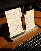 Two note cards with handwritten notes rest in the Analog Card Bar (Small 10" - Silver) solid aluminum card holder on a wooden desk, next to a black pen and in front of a laptop keyboard, illuminated by warm sunlight.