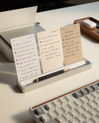 Three notecards featuring handwritten to-do lists are displayed upright on the Analog Card Bar - Essentials Kit (Silver Aluminum), with a black pen resting in front. A keyboard and a wooden tray holding wireless earbuds are also visible on the tidy desk, showcasing the efficient use of dot grid cards for organizing tasks.