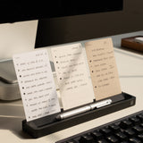 The Analog Card Bar (Small 10" - Black) holds three notecards upright on a desk beside a keyboard and monitor, with a silver pen resting in the holder for an efficient and organized workspace setup.