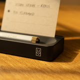 A close-up of the Analog Card Bar (Small 10" - Black), a sleek solid aluminum organizer holding a pen and an index card with handwritten notes. Sunlight creates soft shadows on the black desk accessory atop a wooden surface.