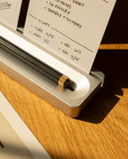 A close-up of the Analog Card Bar - Essentials Kit in Silver Aluminum displays a black pencil with a gold eraser cap resting in the sleek silver holder, surrounded by dot grid cards and notepads featuring handwritten notes and dates, all arranged on a wooden surface illuminated by warm sunlight.