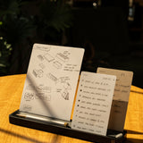 A notepad with hand-drawn sketches and a to-do list rests on a wooden table, held upright by a black stand with a pen. Sunlight shines on the desk, and part of a laptop keyboard is visible in the foreground.