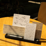 The Analog Card Bar (Small 10" - Black) neatly organizes three notecards with handwritten notes and diagrams, while a pen rests in its tray. Part of a laptop sits nearby on a sunlit wooden table.
