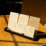 The Analog Card Bar (Small 10" - Black) holds neatly arranged handwritten note cards with to-do lists and notes beside a laptop on a wooden table, all illuminated by warm sunlight.