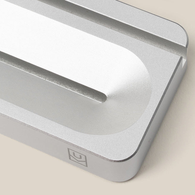 A close-up of a sleek, silver metal object with a slot and a rounded groove, possibly a minimalist desktop accessory or stand, resting on a beige surface.