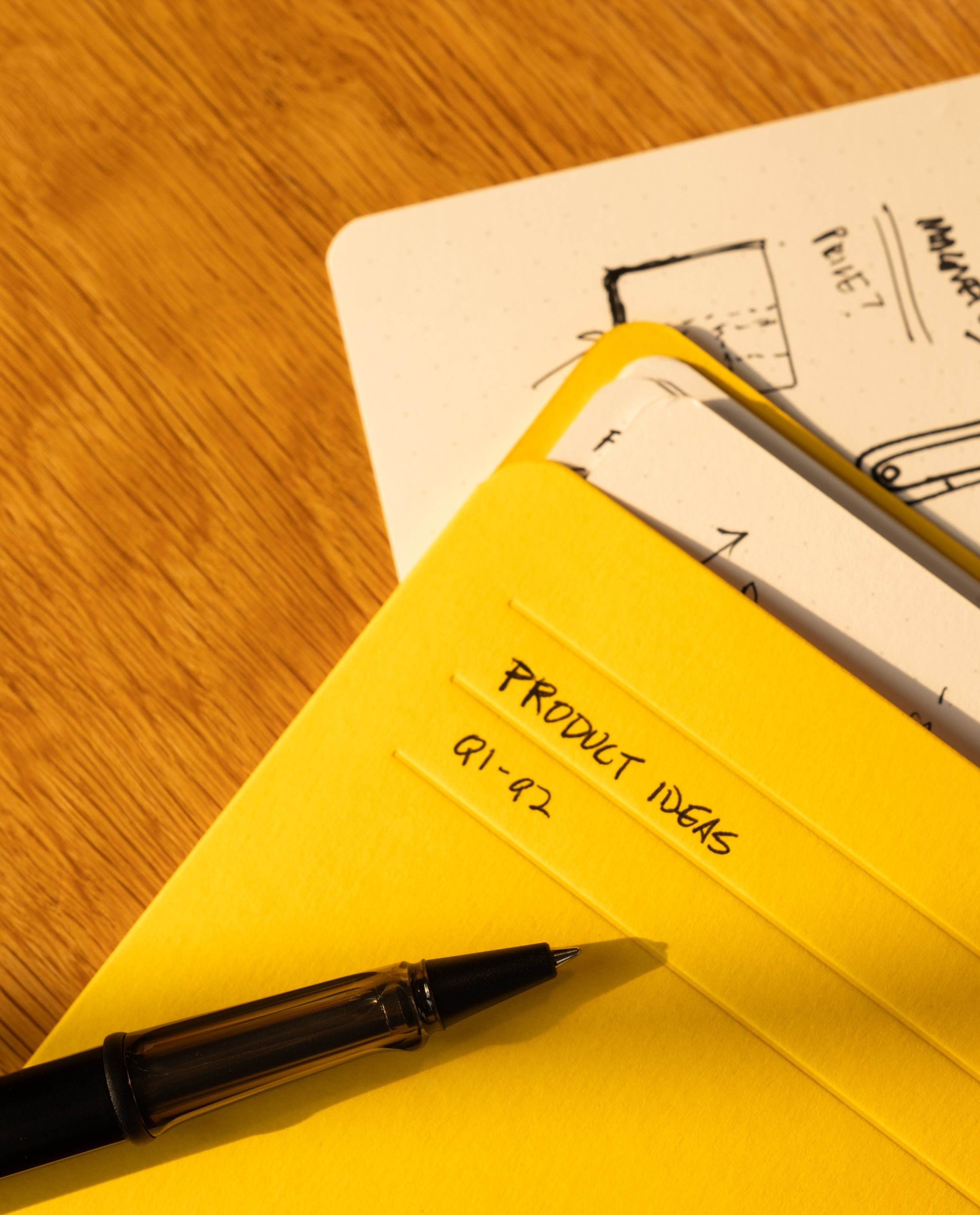 A black pen rests on yellow folders labeled Product Ideas Q1–Q2, beside a dotted notebook filled with sketches and a sleek organizer holding Analog Card Sleeves - Large (Bright). All are arranged on a wooden surface.