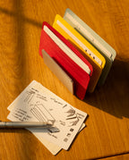 A metal pen rests on a wooden desk beside neatly illustrated note cards, some protected by Analog Card Sleeves - Small (Bright). Behind them, several colorful notebooks stand upright in a metal card organizer, with sunlight casting soft shadows across the scene.