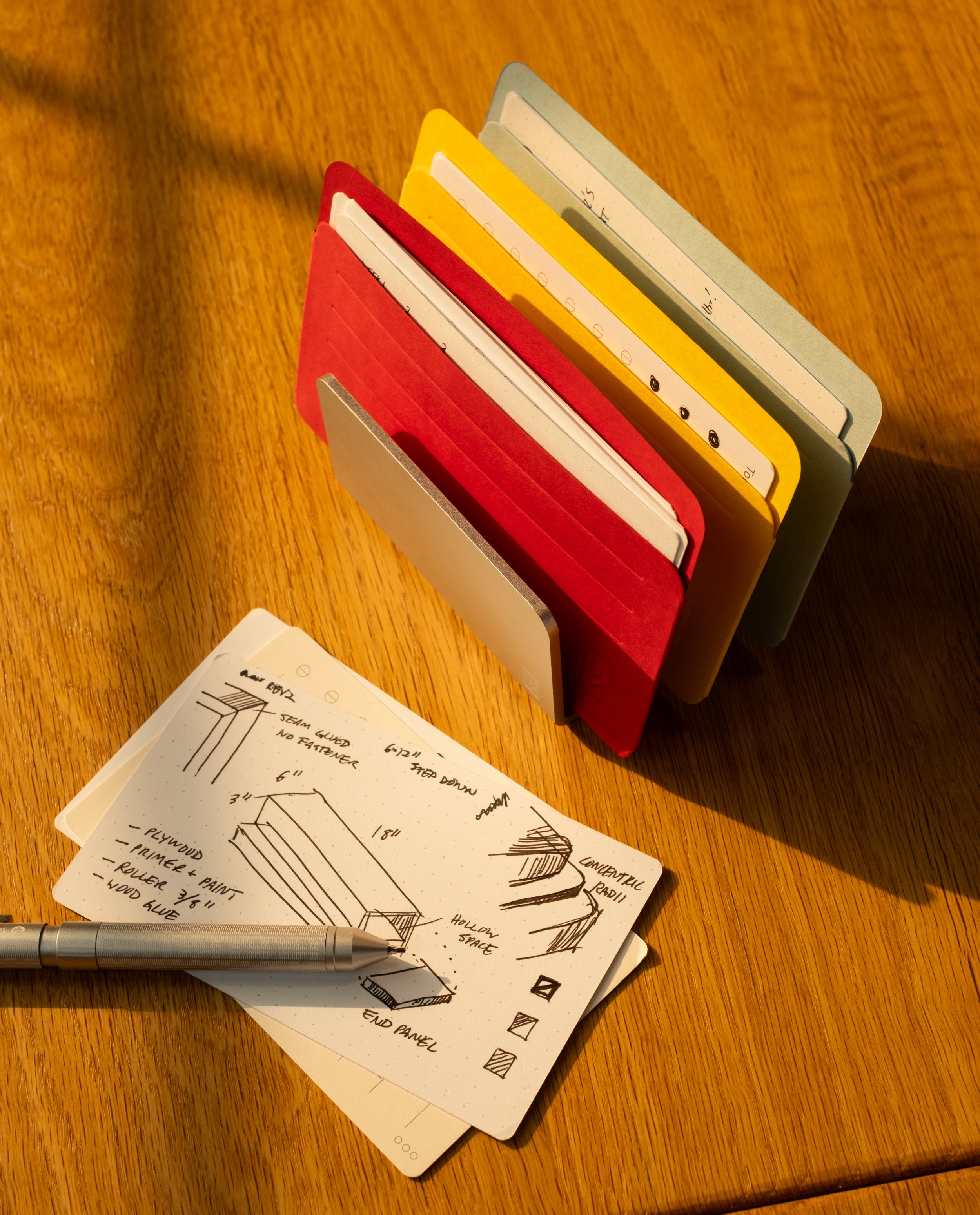 A metal pen rests on a wooden desk beside neatly illustrated note cards, some protected by Analog Card Sleeves - Small (Bright). Behind them, several colorful notebooks stand upright in a metal card organizer, with sunlight casting soft shadows across the scene.