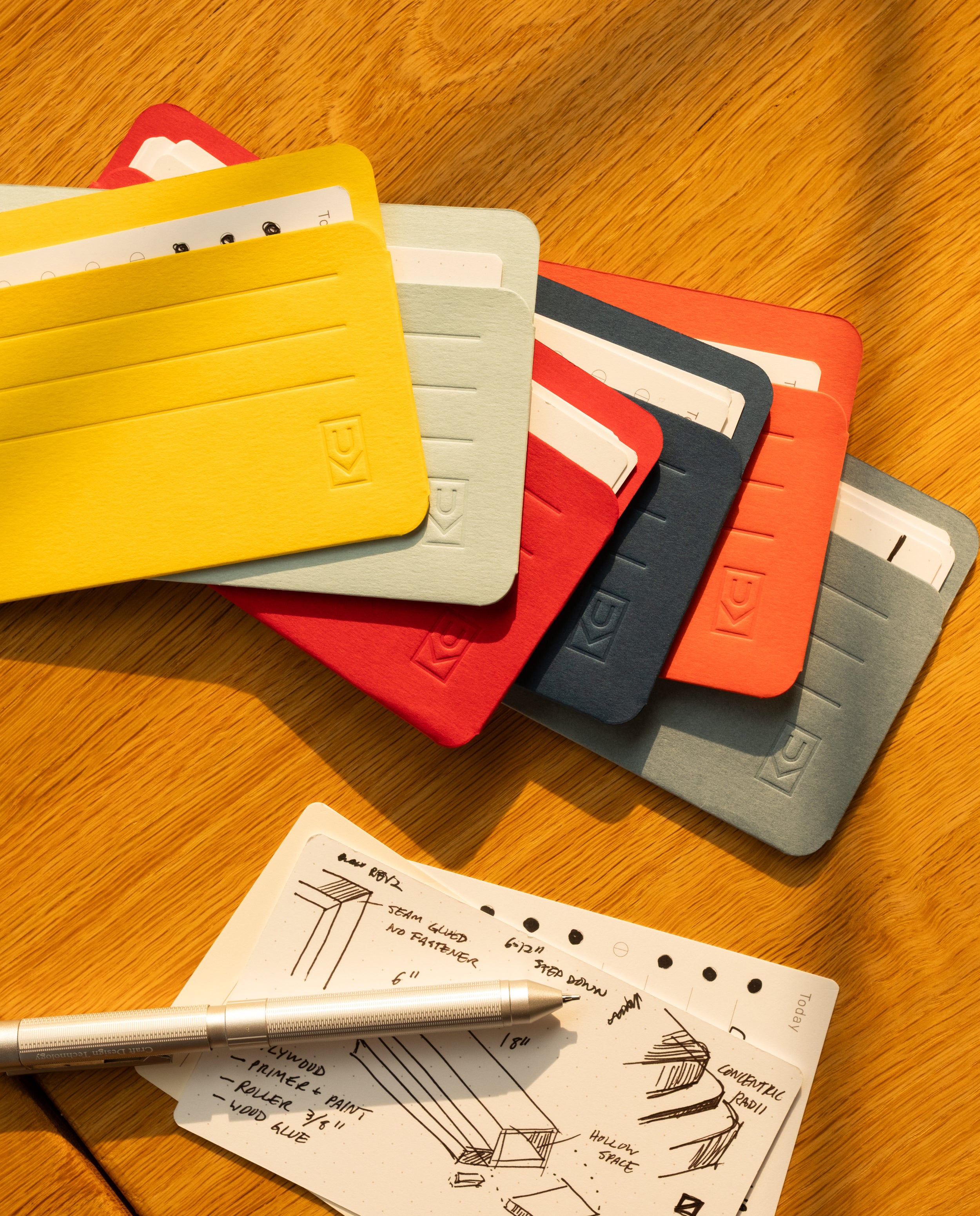 Six vibrant Analog Card Sleeves - Small (Bright) are elegantly fanned out on a wooden table, demonstrating their versatility. Nearby, a sheet of handwritten notes or sketches is accompanied by a silver pen, all bathed in warm sunlight.