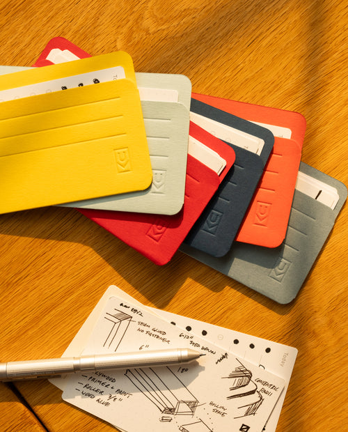 Six vibrant Analog Card Sleeves - Small (Bright) are elegantly fanned out on a wooden table, demonstrating their versatility. Nearby, a sheet of handwritten notes or sketches is accompanied by a silver pen, all bathed in warm sunlight.