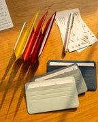 Three Analog Card Sleeves - Small (Bright) and three index cards rest on a wooden desk, creating a simple yet effective card organizer. Nearby are a pen and several small note papers with handwritten notes and doodles. Sunlight casts shadows across the scene, highlighting the charm these bright analog card sleeves bring to an organized workspace.