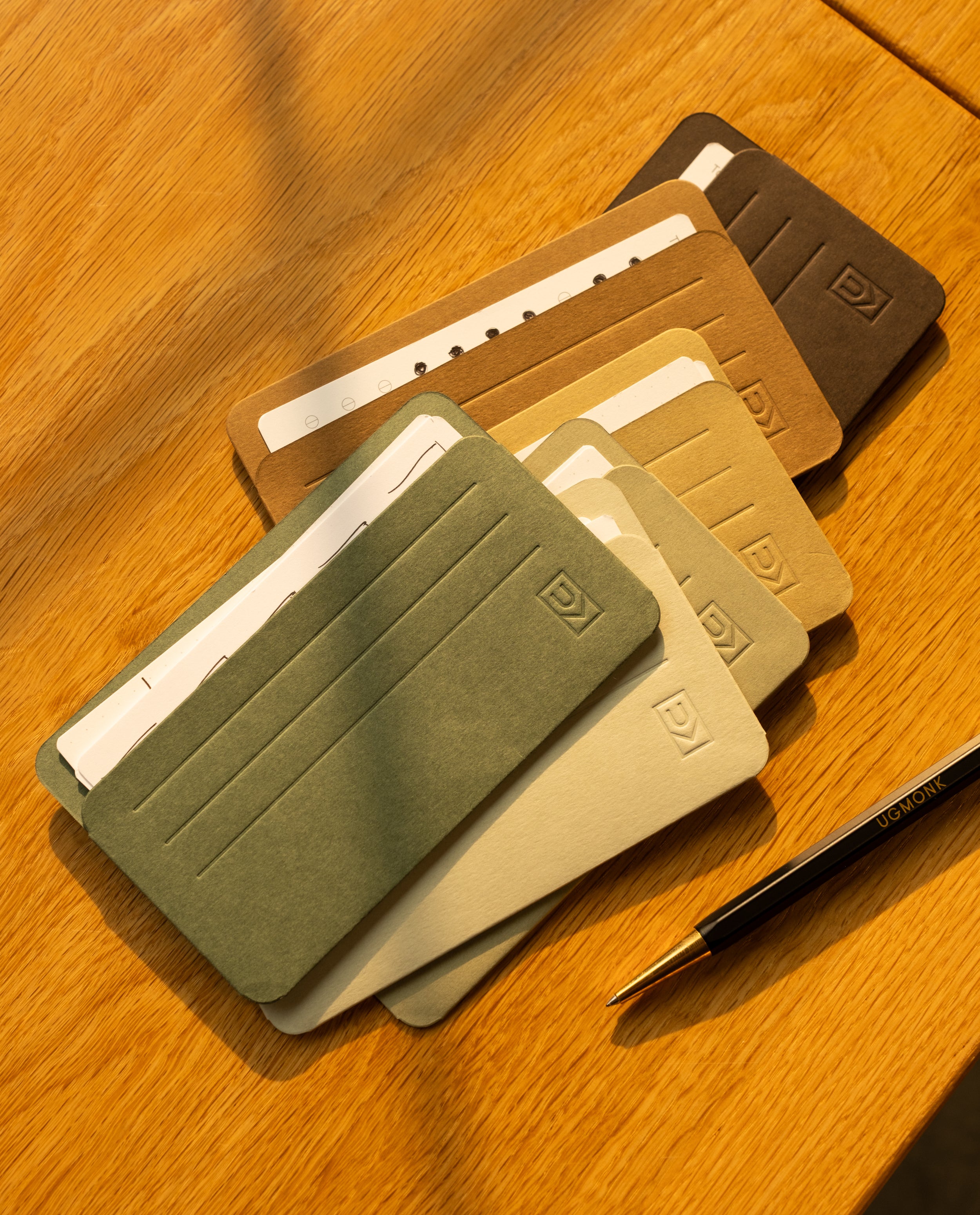 Five slim, minimalist Analog Card Sleeves - Small (Earth) in assorted earth tones are arranged on a wooden surface, each securely holding several cards. A black and gold pen rests nearby as sunlight casts gentle shadows over the scene, accentuating the refined design of these card organizers.