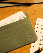 A close-up of green and brown Analog Card Sleeves - Small (Earth) on a wooden surface, with lined and illustrated note cards partially visible underneath—perfect for organizing your notes or pairing with other analog accessories to keep your workspace tidy and accessible.