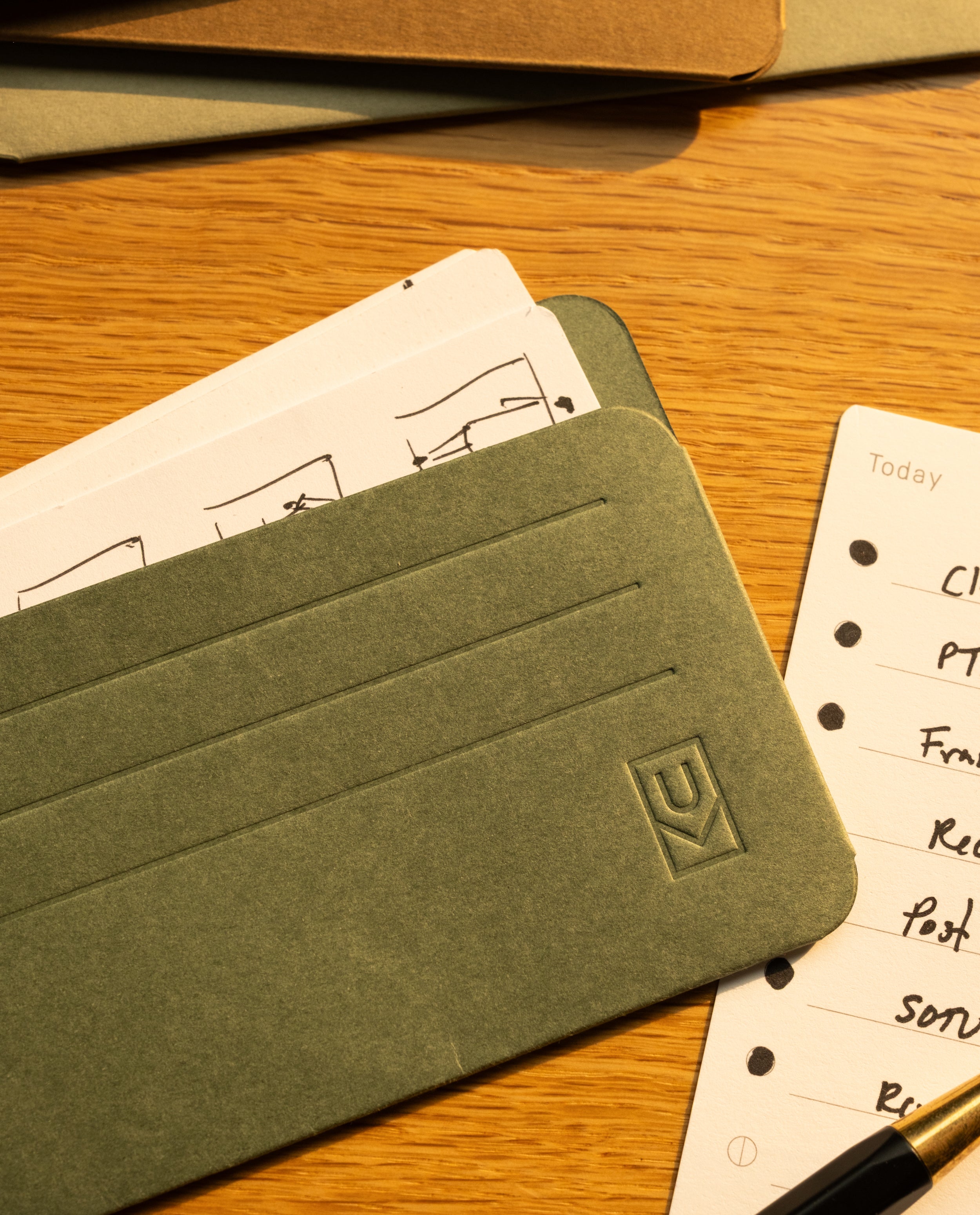 A close-up of green and brown Analog Card Sleeves - Small (Earth) on a wooden surface, with lined and illustrated note cards partially visible underneath—perfect for organizing your notes or pairing with other analog accessories to keep your workspace tidy and accessible.